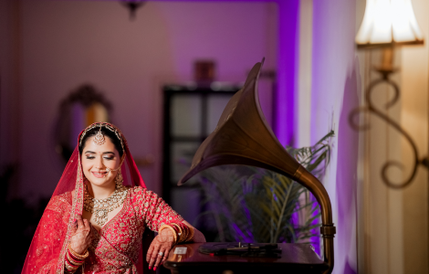In bridal photography, a bride in traditional Indian attire poses at a table with an antique gramophone, exuding grace.