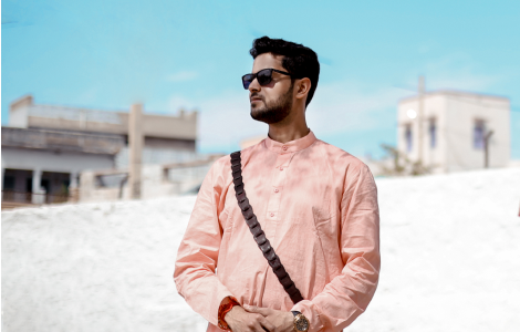A man in a pink traditional kurta and sunglasses stands by a modern building.