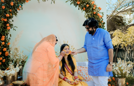 A couple dressed in orange saris sits on a bench, elegantly portrayed in a Haldi Photography style.