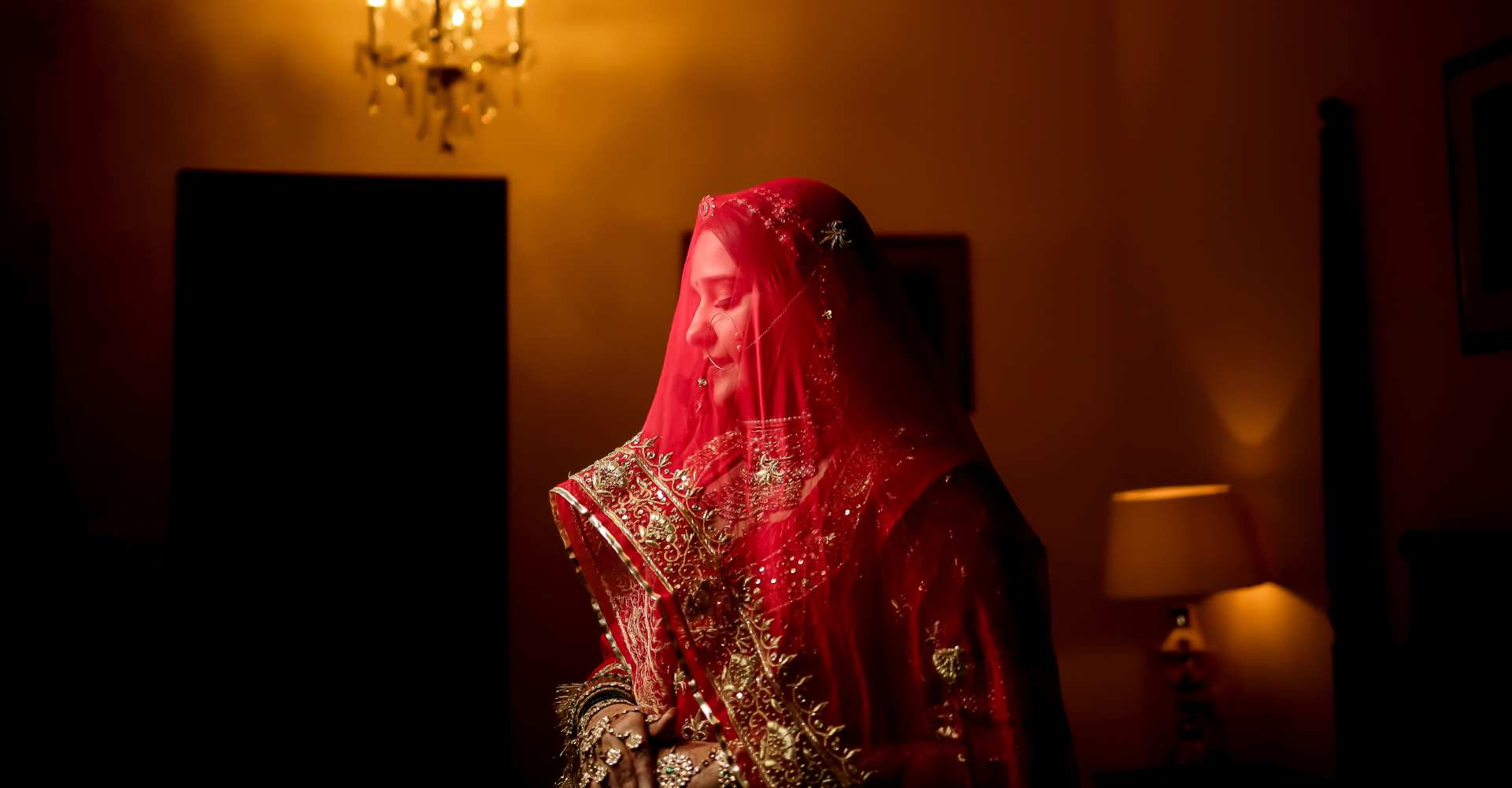 A bride wearing a bold red veil stands in shadows