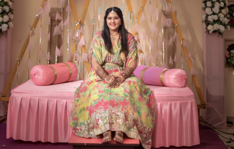 A woman dressed in a colorful outfit sits gracefully on a pink bed, emphasizing her exquisite mehendi artistry.