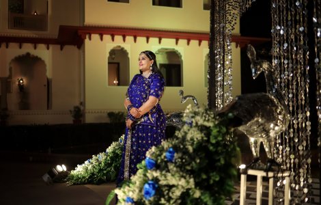 A woman in a blue sari poses gracefully before a vibrant flower arrangement at a wedding sangeet celebration.