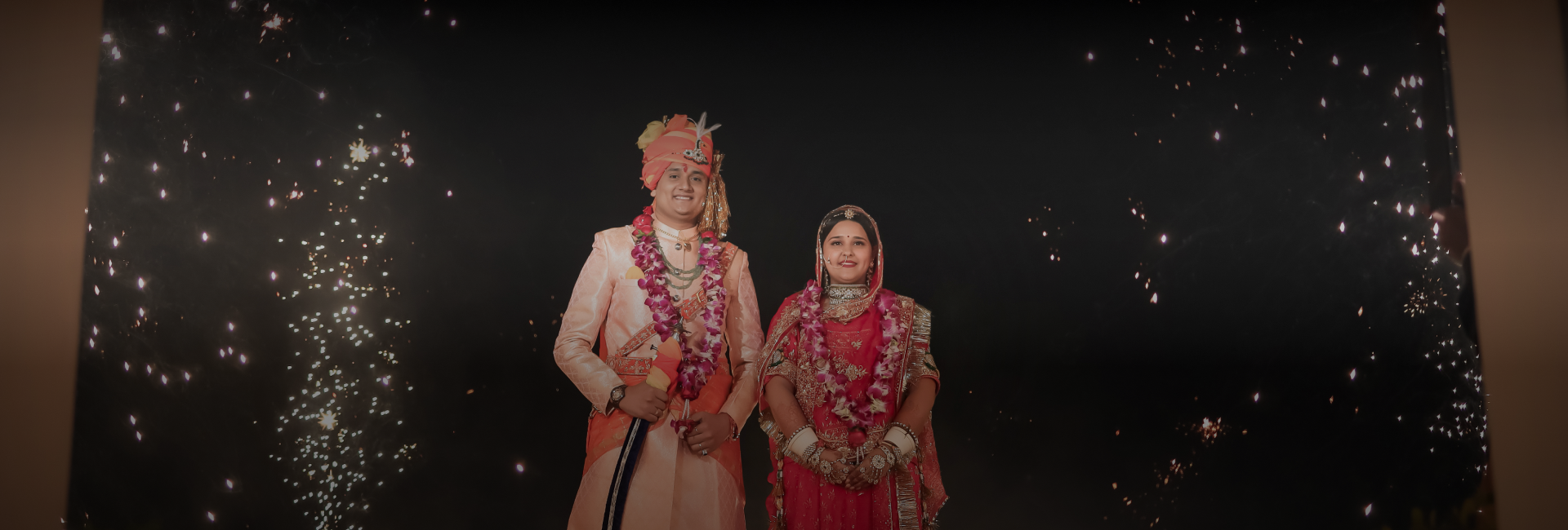 A couple in traditional Indian attire poses joyfully in front of vibrant fireworks, capturing a moment of wedding celebration.